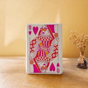 Könnte beinhalten: Eine rechteckige weiße Dose mit einer lebendigen Grafik einer Herz-Dame-Spielkarte. Das Design verwendet kräftiges Pink, Orange und Weiß und zeigt eine stilisierte Figur mit herzförmiger Sonnenbrille, die eine kleine Blume hält.