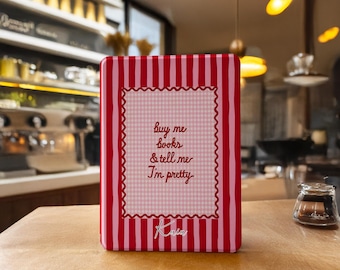 May include: A rectangular, red and pink striped book with the text "buy me books & tell me I'm pretty" in a decorative frame. The book is sitting on a wooden surface.