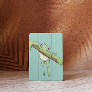 May include: A light blue tablet case featuring a cartoon frog hanging from a mossy branch. The frog has large eyes and a white belly. The branch is brown and green, set against a textured background.
