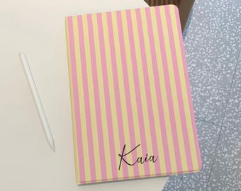 May include: A notebook with a pink and yellow striped pattern. The word "Kaia" is written in cursive on the cover. A white stylus is next to the notebook on a white surface.