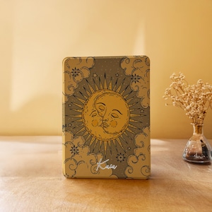May include: A rectangular metal tin featuring a gold and dark gray illustration of a sun and moon face intertwined. The design includes surrounding clouds and stars with the word Kaia written in white script at the bottom center of the surface.