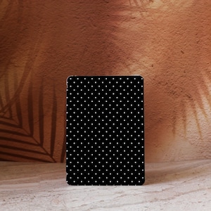 Puede incluir: Un objeto rectangular negro con un patrón de lunares blancos. Se encuentra sobre un fondo marrón texturizado con una sombra de hoja. La superficie sobre la que descansa es de color marrón claro.