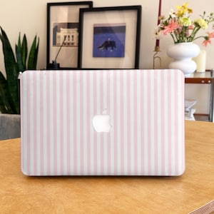 Peut inclure: Un ordinateur portable avec un motif à rayures roses et blanches. Le logo Apple est visible au centre. L'ordinateur est posé sur une table en bois. Des cadres et un vase de fleurs sont en arrière-plan.