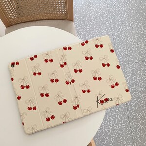 May include: A cream-colored iPad case with a cherry and bow pattern. The case is closed and the name "Kasia" is written on the front.