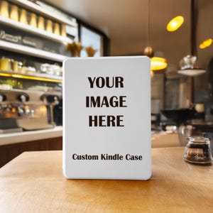 Könnte beinhalten: Eine weiße, rechteckige Kindle-Hülle mit dem Text "YOUR IMAGE HERE" in fetten schwarzen Buchstaben. Darunter steht "Custom Kindle Case" in kleinerer Schrift. Die Hülle steht auf einem Holztisch in einem Café.