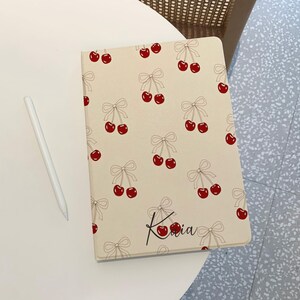 May include: A cream-colored iPad case with a repeating pattern of red cherries and white bows. The name "Karia" is written in black cursive on the front of the case.