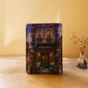 May include: A rectangular metal tin featuring a detailed illustration of a purple-toned book shop exterior with climbing vines, lit windows, and the text "BOOK SHOP" above the entrance and the name "Kaia" written in white script at the bottom.