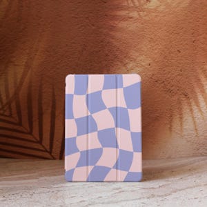 May include: A tablet case with a wavy checkerboard pattern in pastel pink and periwinkle blue. The case is rectangular and has a folding cover. The background is a textured brown surface with a leaf shadow.