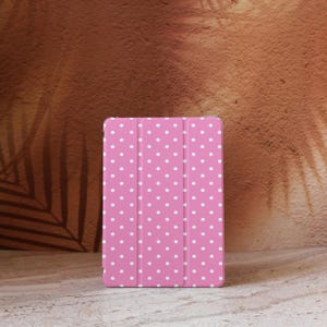 Könnte beinhalten: Eine pinkfarbene Tablethülle mit weißem Polka-Dot-Muster. Die Hülle ist geschlossen und hat ein dreifaches Design. Der Hintergrund ist eine strukturierte braune Oberfläche.