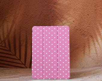 Op de afbeelding: Een roze tablethoesje met een wit stippenpatroon. De hoes is gesloten en heeft een drievoudig ontwerp. De achtergrond is een getextureerd bruin oppervlak.