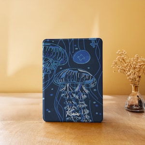 Puede incluir: Una funda azul marino con una ilustración detallada de medusas en tonos azules. El diseño incluye la firma del artista en blanco. La funda es rectangular con esquinas redondeadas.