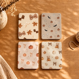 May include: Four rectangular e-reader cases featuring autumn-themed patterns. One case has brown bows and maple leaves, another has ghosts, pumpkins, and bats, the third has pumpkins and ribbons, and the fourth has teddy bears, bows, and leaves.