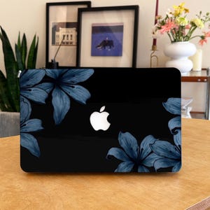 May include: A black laptop skin featuring a dark blue lily floral pattern on the left and right sides. The design frames the centre, where a white Apple logo is clearly visible. The skin has a smooth finish and covers the exterior of the laptop.