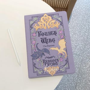 Puede incluir: Una portada de libro de color lila con el título "Fourth Wing" y la autora "Rebecca Yarros". La portada presenta ilustraciones de dragones, detalles florales y filigranas doradas decorativas. Un lápiz óptico blanco descansa sobre la mesa.