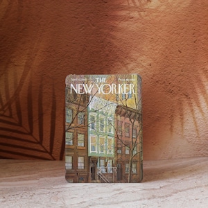 Puede incluir: Una lata rectangular con una colorida ilustración de casas de piedra de Nueva York, con el texto "THE NEW YORKER" en la parte superior. Los edificios son en tonos marrones, verdes y amarillos, con ramas de árboles desnudas en primer plano.