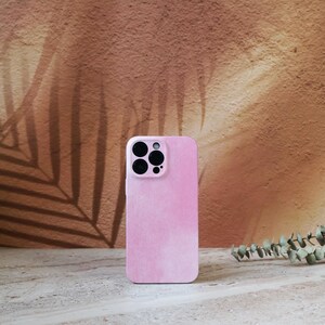 May include: A pink corduroy phone case with a multi-lens camera cutout. The case has a vertical ribbed texture and a soft, pastel colour. The background features a brown wall with a leaf shadow.