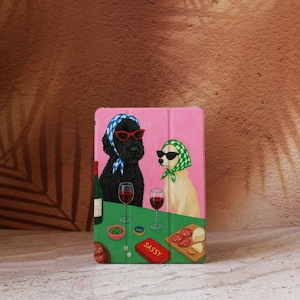 Puede incluir: Funda rosa para tableta con una ilustración caprichosa de dos perros. Uno lleva un pañuelo azul y blanco y gafas de sol rojas, el otro una bufanda verde y blanca y gafas de sol negras. Una botella de vino, copas y aperitivos están sobre la mesa.