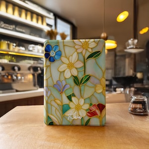 Op de afbeelding: Een rechthoekige hoes met een bloemenontwerp in glas-in-lood. De hoes heeft witte, blauwe, gele en rode bloemen met groene bladeren, omlijnd met goud. De achtergrond is lichtgroen. De naam "Kaia" staat cursief op de hoes.