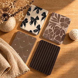 May include: Four rectangular e-reader cases featuring distinct designs: one with black bows on a cream background, one with brown floral patterns, one with a brown grid of white bows and hearts, and one with vertical dark brown and black stripes.