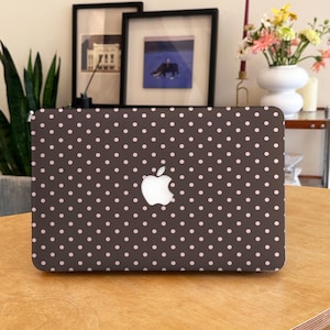 Op de afbeelding: Een laptop met een donkerbruine hoes met een patroon van kleine, gelijkmatig verdeelde roze stippen. Het Apple-logo staat in het midden. De laptop staat op een houten tafel.