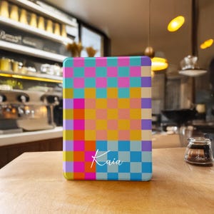 Puede incluir: Una funda rectangular con un colorido patrón de cuadros en rosa, azul, amarillo y naranja. El nombre "Kaia" está escrito en escritura blanca en la parte inferior. La funda está sobre una superficie de madera.