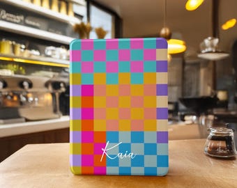 Puede incluir: Una funda rectangular con un colorido patrón de cuadros en rosa, azul, amarillo y naranja. El nombre "Kaia" está escrito en escritura blanca en la parte inferior. La funda está sobre una superficie de madera.