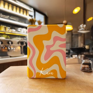 Könnte beinhalten: Eine rechteckige Hülle mit Retro-Design, die wirbelnde Muster in Gelb-, Rosa- und Cremetönen aufweist. Der Name "Kaia" ist in einer Schreibschrift unten geschrieben. Die Hülle liegt auf einer Holzoberfläche.