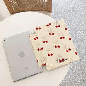 May include: A white iPad case with a cherry and bow pattern. The case has the name "Kavia" written on it. The case is shown with a white iPad.