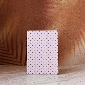 Puede incluir: Una funda para tableta de color rosa claro con un patrón repetido de pequeños lunares marrones oscuros. La funda es rectangular y tiene un diseño plegable triple. El fondo es una superficie marrón texturizada.