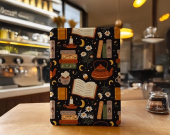 Könnte beinhalten: Eine rechteckige E-Reader-Hülle mit schwarzem Hintergrund und einem wiederkehrenden Muster aus offenen Büchern, Bücherstapeln, einer Teekanne, einer Teetasse, brennenden Kerzen, Mondsicheln und weißen Gänseblümchen. Das Wort Kobo ist in weißer Schrift gedruckt.