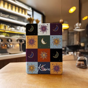 Puede incluir: Una funda rectangular con un diseño celestial de soles, lunas y estrellas en un patrón de cuadrícula. La paleta de colores incluye cuadrados negros, morados, naranjas y azules. El nombre "Kaia" está escrito en cursiva en la parte inferior.