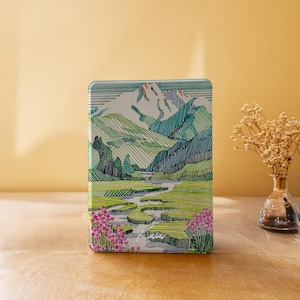 Può includere: Un astuccio rettangolare in metallo con un'illustrazione di paesaggio. L'opera raffigura una catena montuosa con tonalità verdi e blu, un fiume serpeggiante e fiori rosa. L'astuccio ha una finitura lucida e angoli arrotondati.