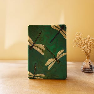 Puede incluir: Una funda rectangular verde con un diseño de libélulas. La funda tiene un fondo verde oscuro con un patrón de remolinos. Varias libélulas con alas color crema y cuerpos marrones están dispersas por la superficie.