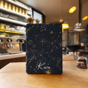 以下が含まれることがあります： 星とつながる線で描かれた、金色の星座のデザインが施された濃い青色のタブレットケース。「Kaia」という名前が白い筆記体で書かれています。ケースは長方形で、木製の表面に置かれています。