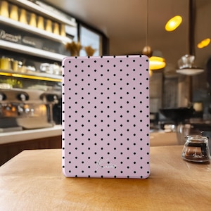 May include: A pale pink tablet case featuring a repeating pattern of black polka dots. The case has a rounded shape and a small signature in the lower right corner. It rests on a wooden surface.