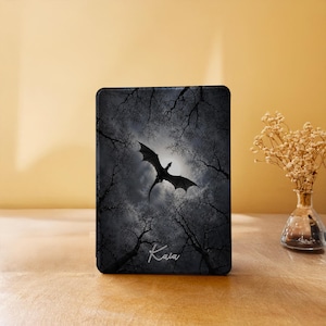 May include: A rectangular e-reader cover featuring a dark, atmospheric forest scene with bare tree branches silhouetted against a bright moon. A black dragon is depicted in flight across the centre. The name "Kara" is written in white script.