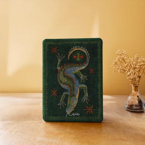 May include: A rectangular dark green notebook featuring a central illustration of a multi-coloured lizard with blue, green, and yellow scales. The cover includes small red star-like symbols and the cursive white text "Kaia" at the bottom.