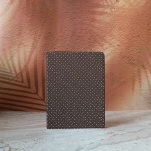 May include: A brown notebook with a white polka dot pattern. The notebook is rectangular and stands upright against a textured, reddish-brown background with a palm leaf shadow.