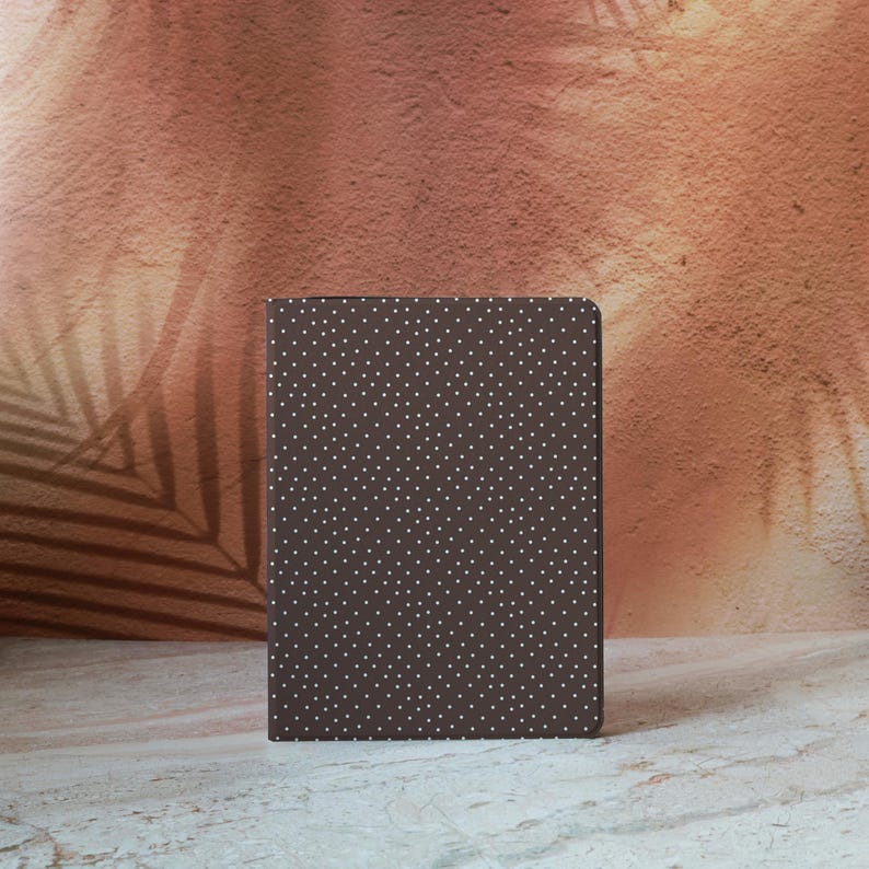 May include: A brown notebook with a white polka dot pattern. The notebook is rectangular and stands upright against a textured, reddish-brown background with a palm leaf shadow.