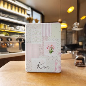 May include: A pink and white patchwork design tin box with a floral pattern and the name Kaia printed on the front.