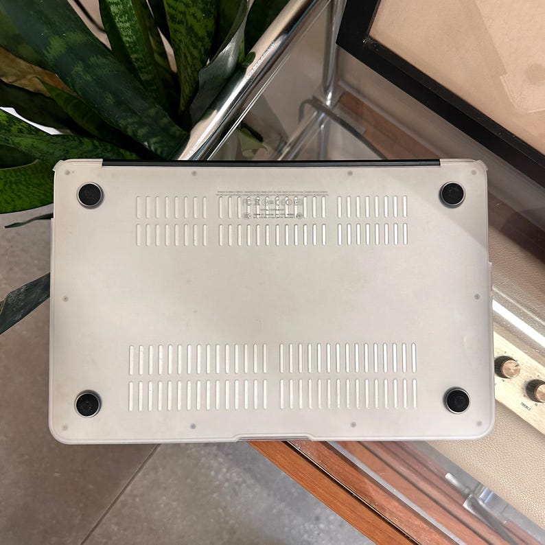 May include: A clear laptop case with a white base. The case is made of a transparent material and has ventilation slots and four black rubber feet. The case is designed to fit a laptop.