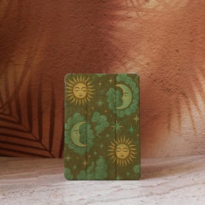 Puede incluir: Una funda verde para tableta con un diseño celestial de soles, lunas, estrellas y nubes. Los soles y las lunas tienen caras, sobre un fondo texturizado. La funda es rectangular con esquinas redondeadas.