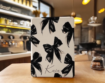 May include: A rectangular white case featuring a repeating pattern of black bows and the name Kaia written in black cursive script. The object has a smooth, flat surface and is positioned upright on a wooden surface.