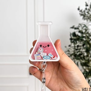 May include: A white and pink badge reel shaped like a laboratory flask, containing pink liquid and science-themed charms. The reel has a smiling face and a retractable cord with a clip.