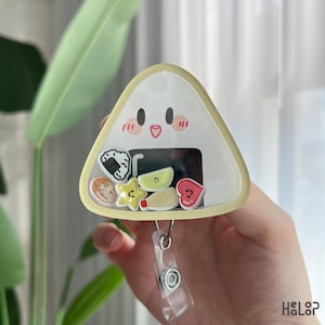 Könnte beinhalten: Ein dreieckiges Ausweis-Rollband mit einem Cartoon-Onigiri-Design und einem lächelnden Gesicht. Die transparente Kunststofffront enthält charms mit Essensmotiven wie Sushi, einem Stern und einem Herz. Das Rollband ist an einem transparenten Kunststoffclip befestigt.