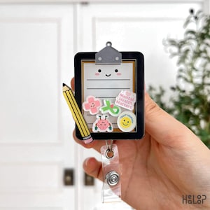 Puede incluir: Un portatarjetas con forma de portapapeles en blanco y negro con un lápiz y varias pegatinas de colores. Las pegatinas incluyen una cruz rosa, una cinta verde, un cerebro y una carita sonriente amarilla con las palabras "Mental Health Matters" y "Trust Yourself".