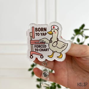 May include: A clear acrylic badge reel with a cartoon duck in a nurse's cap and stethoscope. The badge reel has the text "BORN TO YAP FORCED TO CHART" in pink and white. The duck is white with yellow detailing.