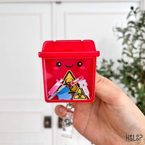 Puede incluir: Un carrete de insignia rojo con un contenedor de riesgo biológico de dibujos animados con una cara sonriente. Dentro del contenedor hay artículos médicos y un símbolo de riesgo biológico. El carrete se sujeta con un clip de plástico transparente.