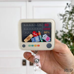 May include: A medical-themed badge reel featuring a monitor screen design with vital signs displayed. The reel is adorned with miniature medical-related charms, including a bandage, heart, syringe, and IV bag. The reel is held by a clear plastic strap.