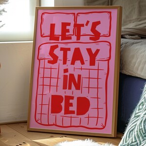 May include: A framed print with the words "LET'S STAY IN BED" in red on a pink background. The artwork has a bed-like design with a grid pattern. The frame is made of light brown wood. The print is leaning against a wall.